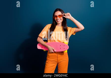 Foto di una donna piacevole vestita di abbigliamento arancione che tocca gli occhiali da sole tenendo il suo nuovo skateboard isolato su sfondo blu scuro Foto Stock