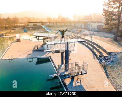 Una persona sta su una piattaforma per immersioni in una piscina all'alba in inverno, nella piscina all'aperto ENCW Calw, Stammheim, Foresta Nera, Germania Foto Stock