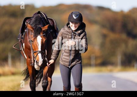 Una giovane donna cammina lungo un sentiero in una soleggiata giornata autunnale e porta il suo cavallo a fare un giro. Vestiti con abiti da equitazione e sellati pronti per il cavallo. Foto nelle terre Foto Stock