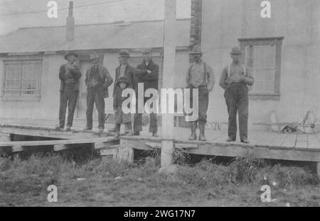 Nativi, tra c1900 e 1916. Ritratto di gruppo di sei uomini e un ragazzo in piedi di fronte a un edificio in latta ondulata, Tanana(?), Alaska. Foto Stock