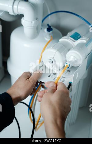 un uomo caucasico, in tute blu, installa un sistema domestico di osmosi inversa sotto il lavandino di una cucina Foto Stock