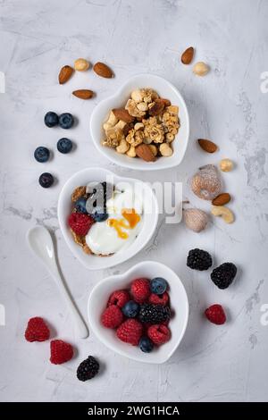 trio di ciotole a forma di cuore disposte su uno sfondo leggermente ruvido con frutti di bosco, frutta secca, yogurt bianco, fiocchi integrali e miele Foto Stock