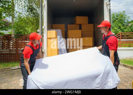 Due uomini che si spostano da un'azienda assistono diligentemente nello scarico di un divano dal veicolo davanti alla casa di un cliente Foto Stock