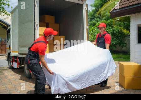 Due uomini che si spostano da un'azienda assistono diligentemente nello scarico di un divano dal veicolo davanti alla casa di un cliente Foto Stock