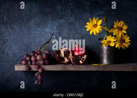 Sulla rustica piattaforma in legno, un mucchio di uva, melograni e fiori gialli di carciofo di Gerusalemme. Natura morta Foto Stock