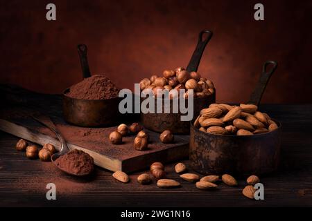 Natura morta. in primo piano con sfondo scuro, misuratori in metallo vintage con nocciole sgusciate, mandorle e cacao in polvere Foto Stock