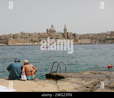 Una coppia di anziani si siede sulle rocce che si affacciano sul mare verso il lungomare della Valletta a Malta in un giorno d'estate. Foto Stock