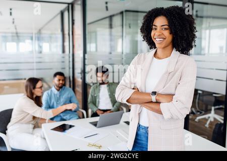 Giovane donna brasiliana intelligente e altamente qualificata, con un sorriso allegro, sta con le braccia incrociate in prima linea in un ambiente di ufficio moderno con i suoi colleghi che collaborano in background, sinergia di squadra Foto Stock