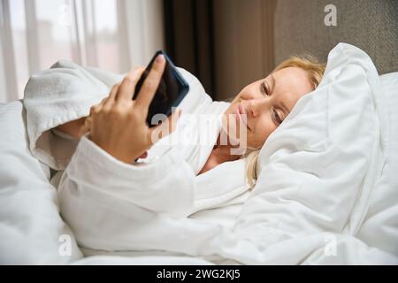 Signora sorridente che usa il telefono cellulare in camera da letto al risveglio mattutino Foto Stock