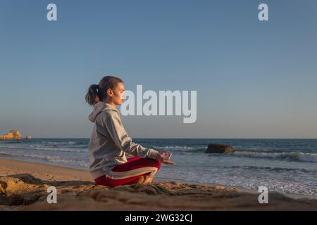 Donna sportiva seduta sulla spiaggia che medita. Surfisti o nuotatori. Foto Stock
