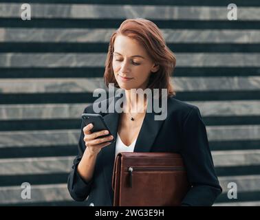 Donna di mezza età con abbigliamento formale nero che guarda il suo smartphone mentre si trova all'aperto Foto Stock