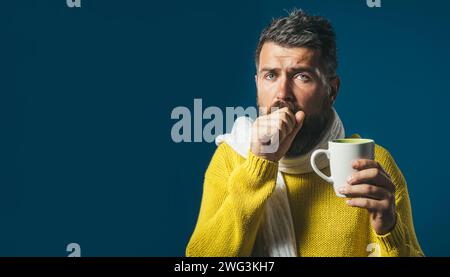 Uomo barbuto malato con influenza o febbre tosse e bere tè curativo. Uomo malvagio che tossisce nel pugno. Uomo malato in maglione avvolto in sciarpa con tazza Foto Stock