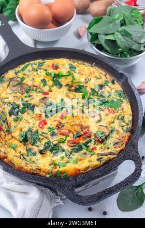 Frittata all'uovo con spinaci, peperoni rossi arrostiti, funghi, formaggio ed erbe aromatiche, in padella di ghisa, verticale Foto Stock