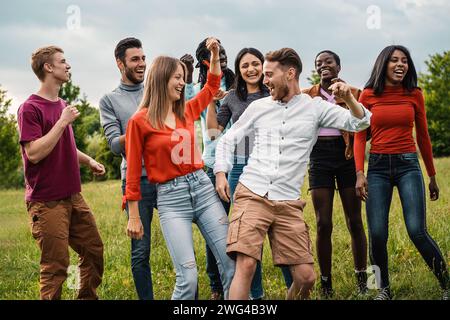 Un vivace gruppo di giovani adulti di varie origini etniche condivide un momento di gioia mentre ballano insieme in un parco, esemplificando lo spirito di un Foto Stock