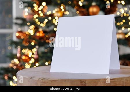 In piedi vuoto, il biglietto d'auguri quadrato vuoto si modella davanti a un albero di natale sfocato con spazio di copia. Da utilizzare come modello di sfondo natalizio. Foto Stock