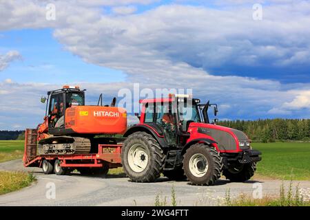 Jokioinen, Finlandia - 13 luglio 2019: Il trattore agricolo Red Valtra T151 trasporta l'escavatore Hitachi Zaxis 130 LCN lungo le strade di campagna nelle giornate di sole dell'estate. Foto Stock