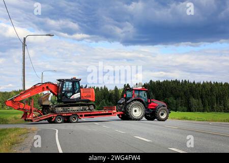 Jokioinen, Finlandia - 13 luglio 2019: Il trattore agricolo Red Valtra T151 trasporta l'escavatore Hitachi Zaxis 130 LCN su un rimorchio dall'altra parte della strada principale del paese Foto Stock