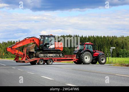 Jokioinen, Finlandia - 13 luglio 2019: Il trattore agricolo Red Valtra T151 trasporta l'escavatore Hitachi Zaxis 130 LCN su un rimorchio dall'altra parte della strada principale del paese Foto Stock