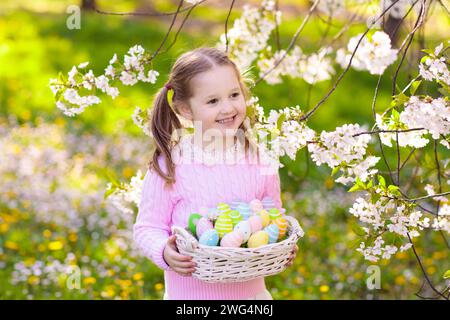 Bambino alla caccia all'uovo di Pasqua in giardino fiorito di ciliegi con fiori primaverili. Bambino con uova colorate nel cestino. Bambina con orecchie conigliate. Foto Stock