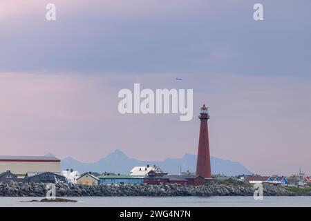 Il faro di Andenes si erge come una sentinella rossa sulla costa rocciosa della Norvegia, con uno sfondo di montagne lontane e un cielo limpido con un piano solitario Foto Stock