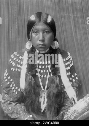 Dusty Dress, c1910. Kalispel Indian Woman, ritratto a metà lunghezza, rivolto verso la parte anteriore, seduta, trecce avvolte, strisce bianche dipinte su capelli, orecchini a disco a conchiglia, indossa un abito coperto decorato con denti di alce. Foto Stock