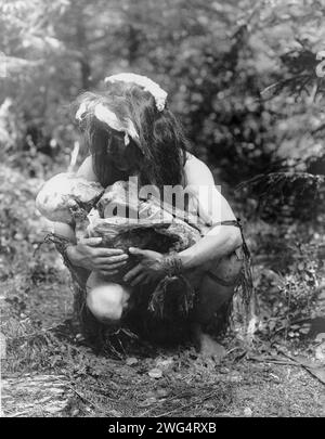 Prepararsi a mangiare la mummia, c1911. Kwakiutl man(?) accovacciata, cullante la mummia legata in posizione fetale. Foto Stock