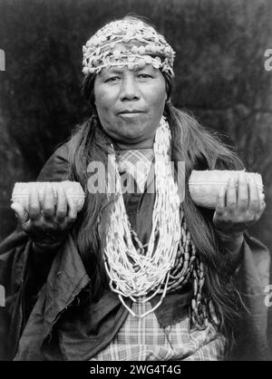 Hupa sciamano femminile, c1923. Athapascan Hupa donna della California nordoccidentale, ritratto a mezza lunghezza, in piedi, davanti, con archetti a conchiglia, collana e tiene in mano due cesti. Foto Stock