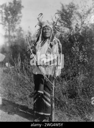 Long Time Dog-Hidatsa, c1908. Long Time Dog, ritratto a tutta lunghezza, in piedi, rivolto verso la parte anteriore, con poggiatesta, camicia in pelle di buccia e leggings con campane in ottone. Foto Stock