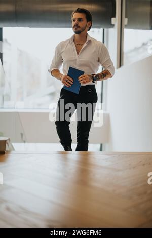 Un giovane ed elegante imprenditore maschio cammina con fiducia in un ufficio, con un notebook blu. Concentratevi sulla determinazione e la professionalità. Foto Stock