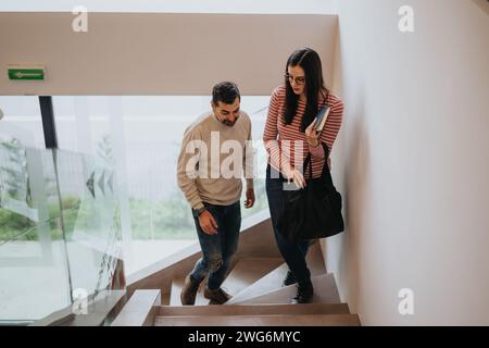 Un giovane uomo e una donna scendono le scale fianco a fianco in un ambiente interno, riflettendo un momento della vita quotidiana a casa o in un ufficio moderno. Foto Stock