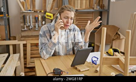 Una giovane donna preoccupata che parla al telefono in un'officina di falegnameria disordinata. Foto Stock