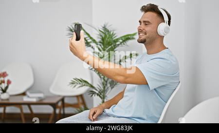 Uomo ispanico sorridente con barba che scatta un selfie in una stanza bianca indossando le cuffie. Foto Stock