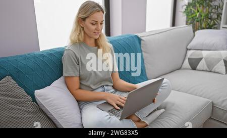 Una giovane donna bionda si rilassa a casa, navigando su un notebook nel suo soggiorno, trasudando comfort e tranquillità. Foto Stock