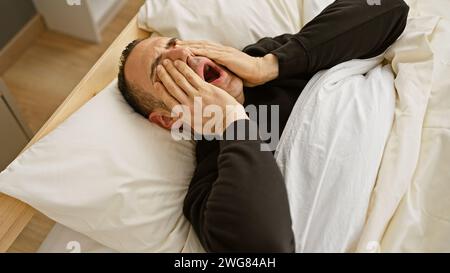 Un giovane angosciato sdraiato a letto in casa si copre il viso con le mani, mostrando segni di tristezza o stanchezza. Foto Stock