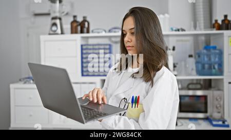 Giovane donna ispanica focalizzata, una bella scienziata al lavoro, che utilizza laptop e microscopio per la ricerca medica in laboratorio, mostrando un rilassato, ma degrado Foto Stock