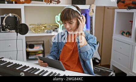 Un giovane ascolta la musica con le cuffie mentre tiene in mano un tablet in uno studio domestico circondato da strumenti musicali. Foto Stock