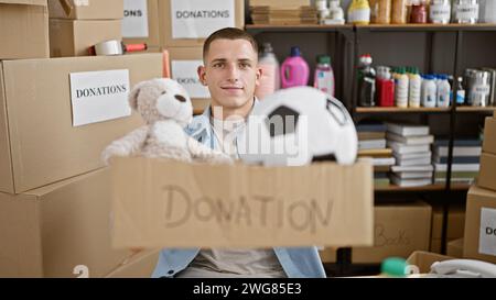 Un giovane sorridente che tiene in mano una scatola per donazioni in un deposito pieno di vari oggetti donati. Foto Stock