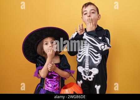 Fratello e sorella che indossano il costume di halloween coprendo la bocca con la mano, scioccati e spaventati per errore. espressione sorpresa su dorso giallo isolato Foto Stock