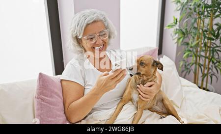 Una donna matura e gioiosa che utilizza uno smartphone coccola il suo cane su un letto all'interno di una stanza accogliente. Foto Stock