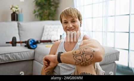 Bel tatuato che sorride in un accogliente soggiorno con attrezzature per l'allenamento sullo sfondo. Foto Stock