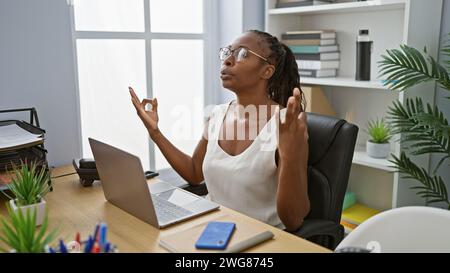 Una giovane donna afroamericana serena medita alla scrivania del suo ufficio circondata da tecnologia e cancelleria. Foto Stock