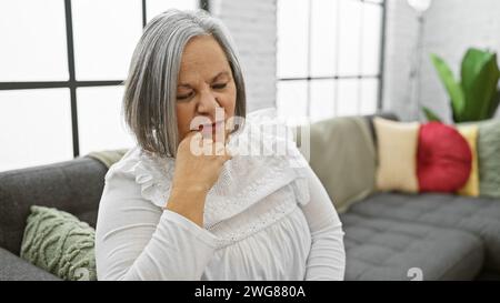 Una donna dai capelli grigi seduto con cura in un salotto accogliente e moderno, riflettendo sull'età adulta matura. Foto Stock
