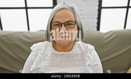 Una donna anziana con capelli e occhiali grigi si siede comodamente a casa su un divano, trasudando calore e maturità. Foto Stock