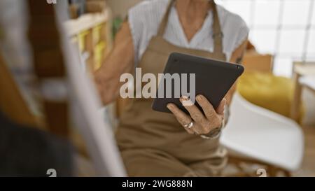Artista anziana sicura di sé, nel suo accogliente studio d'arte, una donna matura disegna piacevolmente su un touchpad circondato da pennelli, tavolozza, cavalletto e tela. Foto Stock