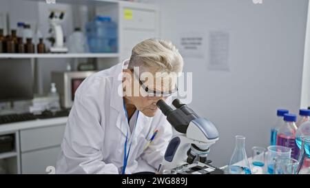 Scienziata senior con i capelli grigi dedicata immersa in un intenso lavoro di ricerca sul microscopio a livello di dottorato in laboratorio Foto Stock