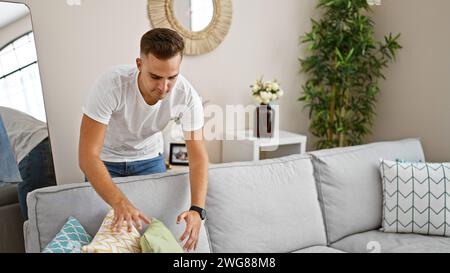 Un giovane uomo dispone di cuscini su un divano in un accogliente e ben illuminato soggiorno, che mostra la vita quotidiana a casa. Foto Stock