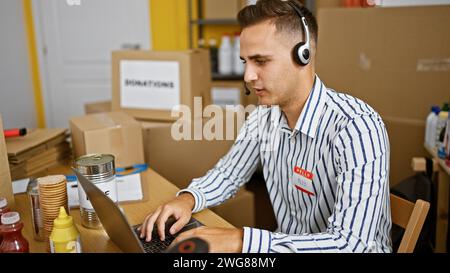 Bel giovane con cuffie che lavora su un computer portatile in un ufficio di magazzino circondato da scatole e forniture alimentari. Foto Stock