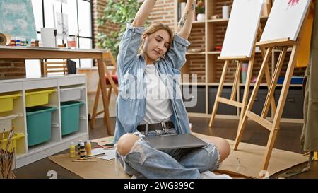 Giovane artista donna che allunga le braccia seduta sul pavimento presso lo studio d'arte Foto Stock