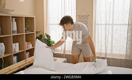 Bel giovane ispanico in piedi, che si concentra a fare il suo letto in una camera da letto illuminata dal sole, trasuda uno stile di vita rilassato tra cuscini e b Foto Stock