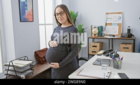 Una donna ispanica incinta si trova con fiducia in un ufficio, catturando temi di maternità e professionalità. Foto Stock